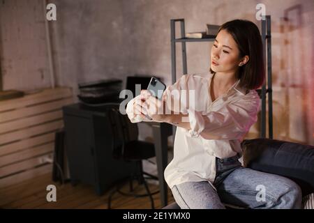 Jeune femme asiatique assise sur un canapé dans un confortable la chambre prend un selfie et sourit Banque D'Images