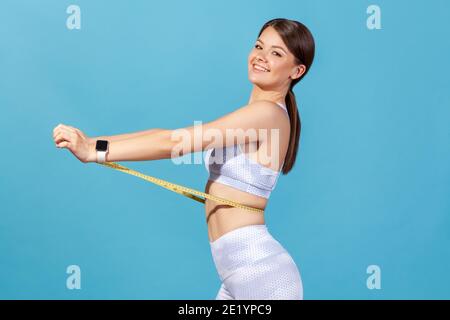 Portrait de profil bonne femme souriante en blanc sportswear mesurer sa taille avec mètre ruban et sourire regardant l'appareil photo, satisfait de l'amincissement. Banque D'Images