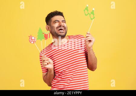 Un homme plein de joie avec une barbe en t-shirt rayé riant fort tenant les mains masques en papier de vacances sur bâton, célébrant Noël et nouvel an. Po Banque D'Images