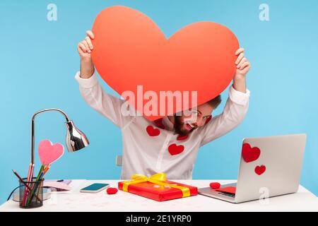 Drôle positif barbu homme peeping out gros coeur de papier rouge qu'il tient, préparant la carte de voeux, assis sur le lieu de travail tous couverts de coeurs collants. JE Banque D'Images