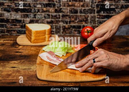 Les mains caucasiennes mâles coupent du bacon avec un couteau de chef sur une planche à découper et préparent les ingrédients pour un sandwich BLT. Banque D'Images