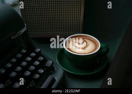 Tasse de café blanc plat dans une tasse verte à côté d'une vieille machine à écrire sur une table en bois vert. Vue rapprochée. Banque D'Images