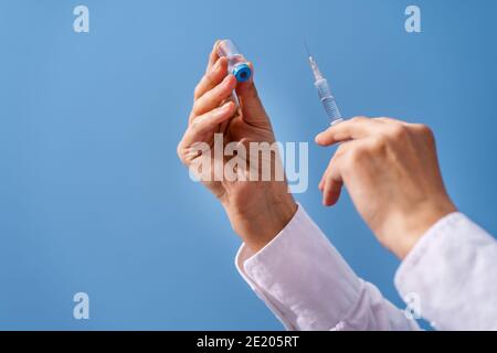 Gros plan sur la main de la femme caucasienne inconnue médecin tenant seringue avec aiguille et injection d'ampoule de vaccin devant paroi bleue - covid-19 coronavir Banque D'Images