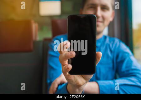 Un gars dans une chemise bleue tient un téléphone portable avec un écran noir pour le texte et le design pendant un trajet en train. Banque D'Images