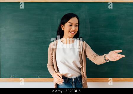 Gaie jeune femme asiatique se gesticuler et regarder loin en se tenant près du tableau noir pendant la leçon à l'école Banque D'Images