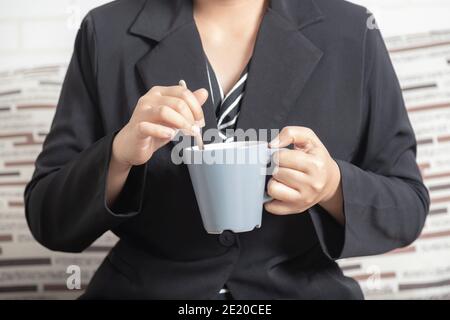 Femme d'affaires en gros plan main tenant une tasse de café et remuant. Femme portant un costume assis sur le canapé pour une pause avec un verre. Banque D'Images