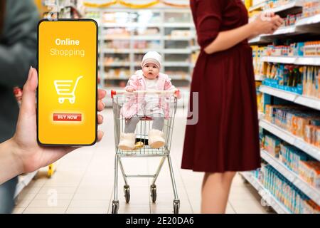 Shopping. Bébé mignon assis dans un panier dans le supermarché, à côté de sa mère. La main gauche contient un smartphone. Le concept de onlin Banque D'Images