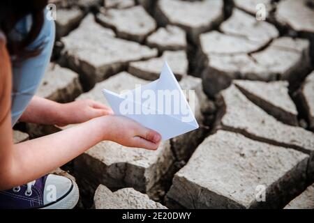 La fille abaisse le bateau en papier sur le sol sec et fissuré. Crise de l'eau et changement climatique concept. Réchauffement de la planète Banque D'Images