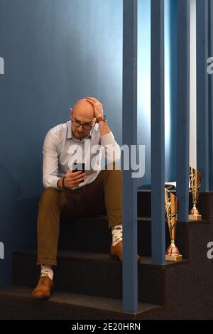 homme d'âge moyen sérieux avec un regard perplexe lisant un message ou un article d'actualité sur son téléphone portable, tenant votre tête Banque D'Images