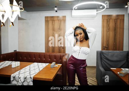 Belle femme afro-américaine en blouse blanche et pantalon en cuir rouge pose au restaurant. Banque D'Images