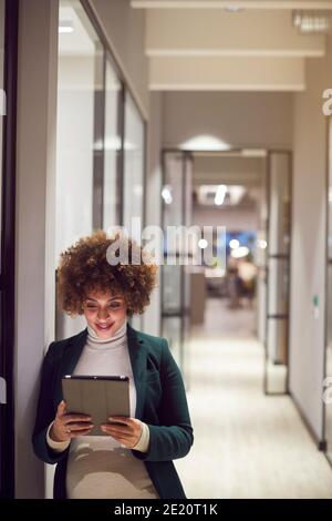 Prise de vue en soirée d'une femme d'affaires enceinte debout dans le couloir du moderne Bureau utilisant une tablette numérique Banque D'Images