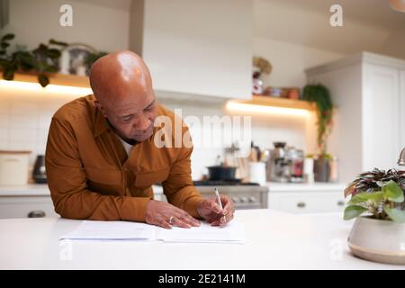 Homme mature qui passe en revue et signant les documents de finances et d'investissement nationaux Dans la cuisine à la maison Banque D'Images