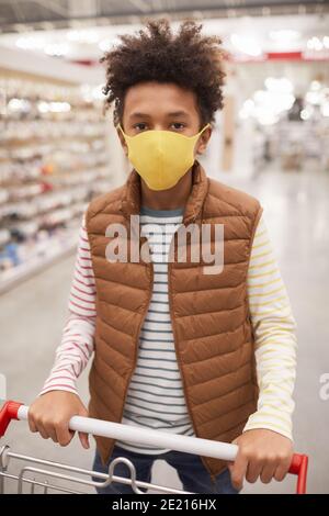 Portrait vertical d'un garçon afro-américain portant un masque dans le supermarché et regarder l'appareil photo tout en poussant le chariot Banque D'Images