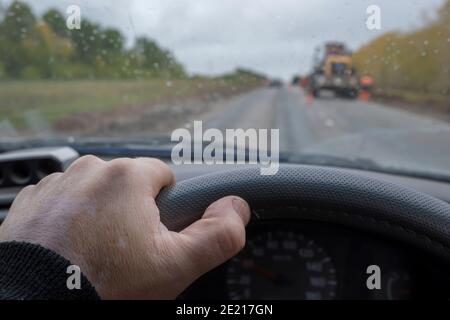 la main du conducteur sur le volant d'une voiture passant devant une section de route en réparation avec un tracteur, équipement de construction par temps pluvieux et nuageux Banque D'Images