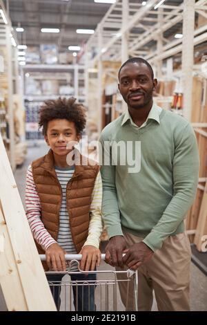 Portrait vertical du père et du fils afro-américains qui magasinent ensemble le matériel de fixation stocke les deux appareils en regardant la caméra tout en poussant le chariot avec planches en bois Banque D'Images