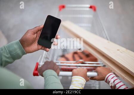 Gros plan d'un homme afro-américain méconnu tenant un smartphone avec un écran vierge tout en poussant le panier dans un supermarché, espace de copie Banque D'Images