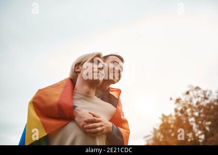 Couple gay romantique embrassant, embrassant et tenant les mains à l'extérieur. Deux hommes séduisant portant un drapeau de fierté LGBT. Banque D'Images