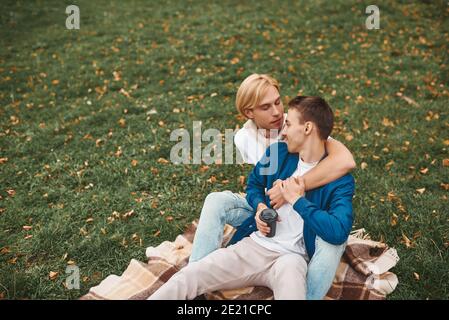 Couple gay amoureux ayant une date romantique à l'extérieur. Deux beaux hommes assis ensemble sur une couverture dans le parc et embrassant. Concept LGBT. Banque D'Images