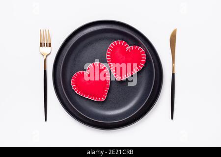 Assiette en céramique noire, fourchette et couteau, coeurs en textile rouge sur fond blanc. Table de Saint-Valentin. Vue de dessus - image Banque D'Images