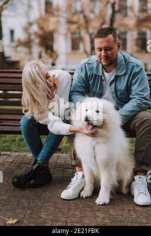 Belle femme et homme apprécient leur date en s'asseyant sur un banc de ruelle avec chien de race samoyed à fourrure blanche Banque D'Images