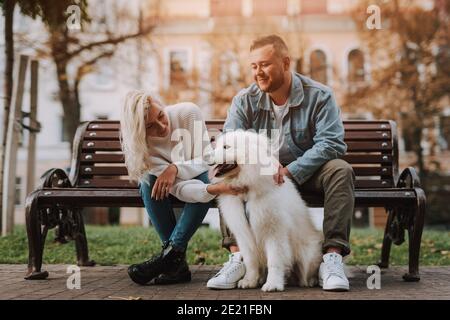 Les jeunes sont des femmes et des hommes qui apprécient la date pendant qu'ils sont assis sur un banc d'allée avec chien à fourrure blanche de race samoyed Banque D'Images