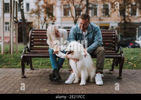 Jeune jolie dame et son petit ami apprécient leur date assis sur un banc d'allée avec un chiot à fourrure blanche race de samoyed Banque D'Images