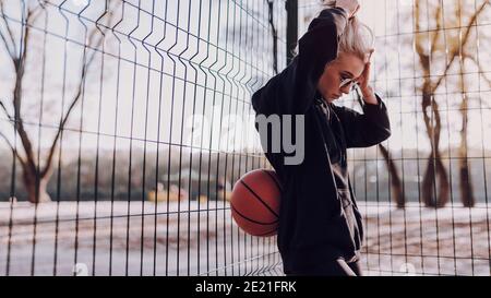 Jeune jolie femme se reposant après avoir joué au basket-ball tout en étant debout à l'extérieur Banque D'Images