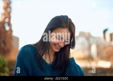 Jeune belle femme regardant vers le bas avec le sourire heureux tout en se fatiguant en haut de ses cheveux avec la main sur fond de ville défoqué rue Banque D'Images