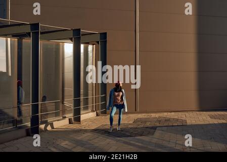Belle jeune femme brunette dans un Jean tendance tenue debout près à un bâtiment moderne de la ville et regardant de côté Banque D'Images