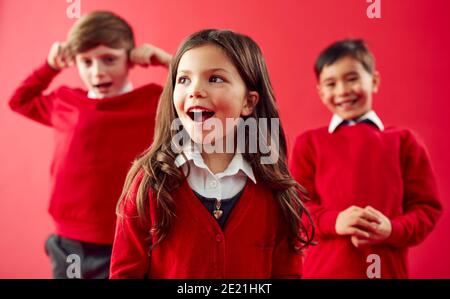 Groupe d'élèves enthousiastes de l'école élémentaire portant un uniforme ayant du plaisir Sur fond Red Studio Banque D'Images