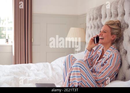 Femme d'affaires en pyjama assise sur le lit faire appel sur Mobile Téléphone à domicile Banque D'Images