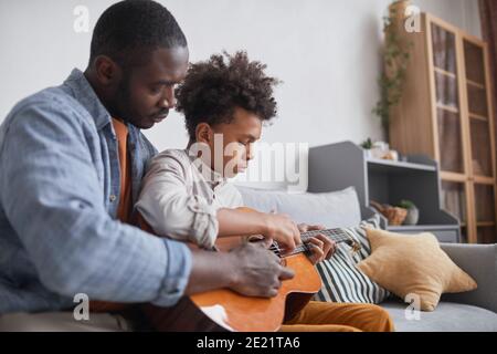 Homme américain moderne d'Afrique passant du temps libre avec son adolescent fils à la maison lui enseignant comment jouer de la guitare Banque D'Images