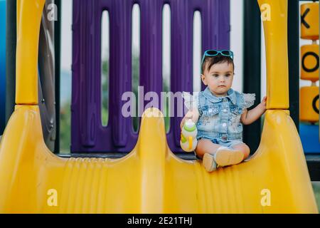 Joli bébé de neuf mois assis sur un toboggan dans l'aire de jeux et tenant son jouet sucette Banque D'Images