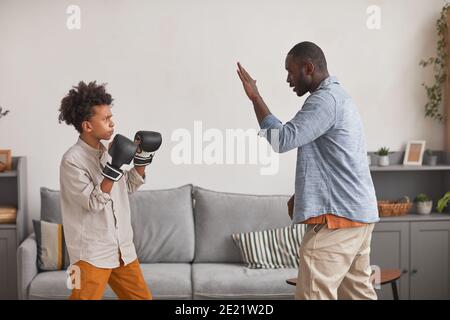 Homme afro-américain sportif pratiquant la boxe avec son fils adolescent dans le salon à la maison, vue latérale Banque D'Images