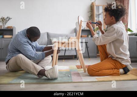 Homme afro-américain moderne passant du temps avec son fils adolescent dans la salle de séjour à la maison, assemblage de la nouvelle chaise Banque D'Images