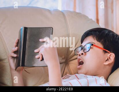 Mignon petit enfant dans des vêtements décontractés lisant un livre et bâilling tout en étant allongé sur le canapé pour apprendre et étudier avec bonheur. Banque D'Images