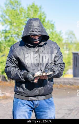 un bandit dans un masque noir se trouve dans la rue et compte l'argent volé Banque D'Images