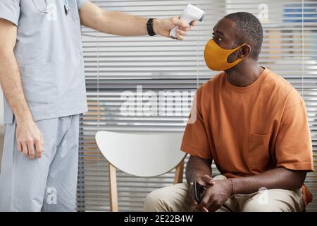 Portrait d'un médecin masculin méconnaissable contrôlant la température de l'homme afro-américain porter un masque en attendant dans la file d'attente à la clinique médicale Banque D'Images