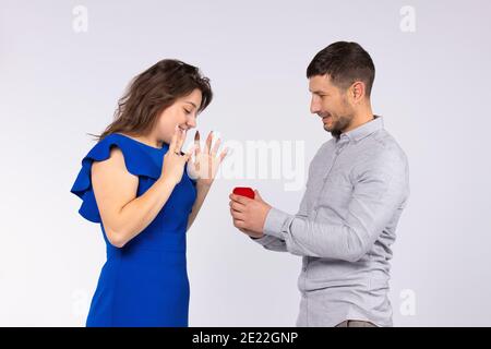 Une fille surprise dans la robe bleue élégante regarde la boîte rouge anneau qu'un gars lui donne. Arrière-plan blanc. Banque D'Images