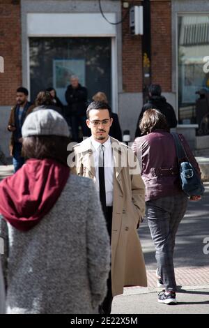 Jeune homme traversant la rue vêtu d'un imperméable, d'une chemise blanche et d'une cravate noire tandis que d'autres personnes sont hors de foyer. Concept de style de vie. Banque D'Images