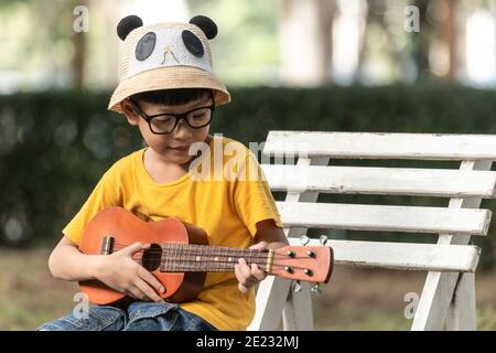 Un petit garçon asiatique portant des lunettes est heureux de jouer l'ukulele. Le petit enfant asiatique essaie de jouer à l'ukulele avec un moment pleinement heureux. Banque D'Images