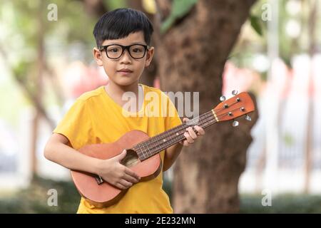Un petit garçon asiatique portant des lunettes est heureux de jouer l'ukulele. Le petit enfant asiatique essaie de jouer à l'ukulele avec un moment pleinement heureux. Banque D'Images
