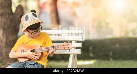 Un petit garçon asiatique portant des lunettes est heureux de jouer l'ukulele. Le petit enfant asiatique essaie de jouer à l'ukulele avec un moment pleinement heureux. Banque D'Images