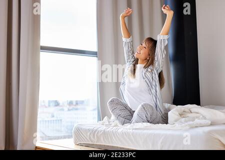 femme asiatique étirant les bras après s'être réveillée le matin, assise sur le lit avec les bras levés, sourire Banque D'Images