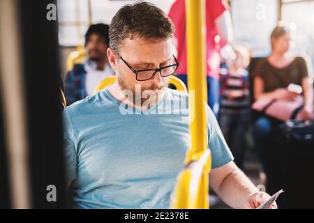 Sérieux homme mature avec des lunettes assis dans le bus plein de gens et de lecture de journaux. Banque D'Images