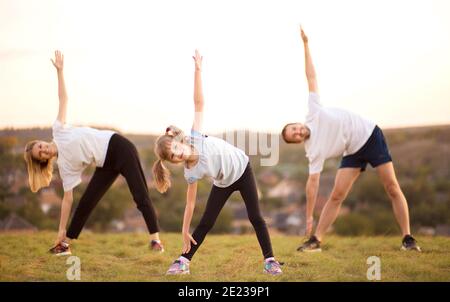 Les parents inculquent des habitudes sportives à leur enfant par exemple, se concentrent sur l'enfant. Bonne famille sportive faisant des exercices d'étirement, pratique le yoga en plein air. Maman papa et fille faisant des exercices de sport ensemble. Banque D'Images