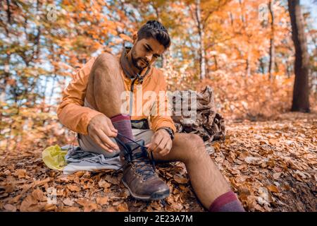 Un randonneur nouant des lacets tout en étant assis sur le sol. À côté de lui sac à dos. Bois à l'extérieur de l'automne, arbres et feuilles mortes tout autour. Banque D'Images