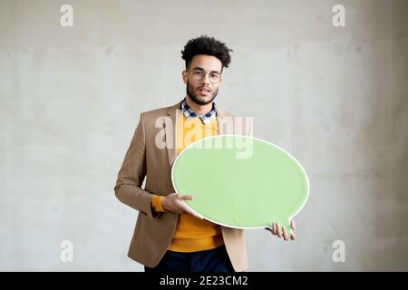 Jeune homme d'affaires élégant de course mixte vous regarde tout en vous tenant debout avant de l'appareil photo et tenant une bulle d'expression verte poussiéreuse Banque D'Images