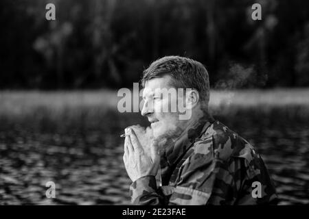 Pêcheur en camouflage fumant une cigarette sur un bateau en caoutchouc dans le lac. Photo en noir et blanc. Banque D'Images