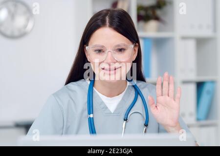 Joyeux jeune brunette médecin en uniforme et lunettes salutation en ligne patient en agitant la main tout en étant assis devant l'ordinateur portable dans le bureau médical Banque D'Images